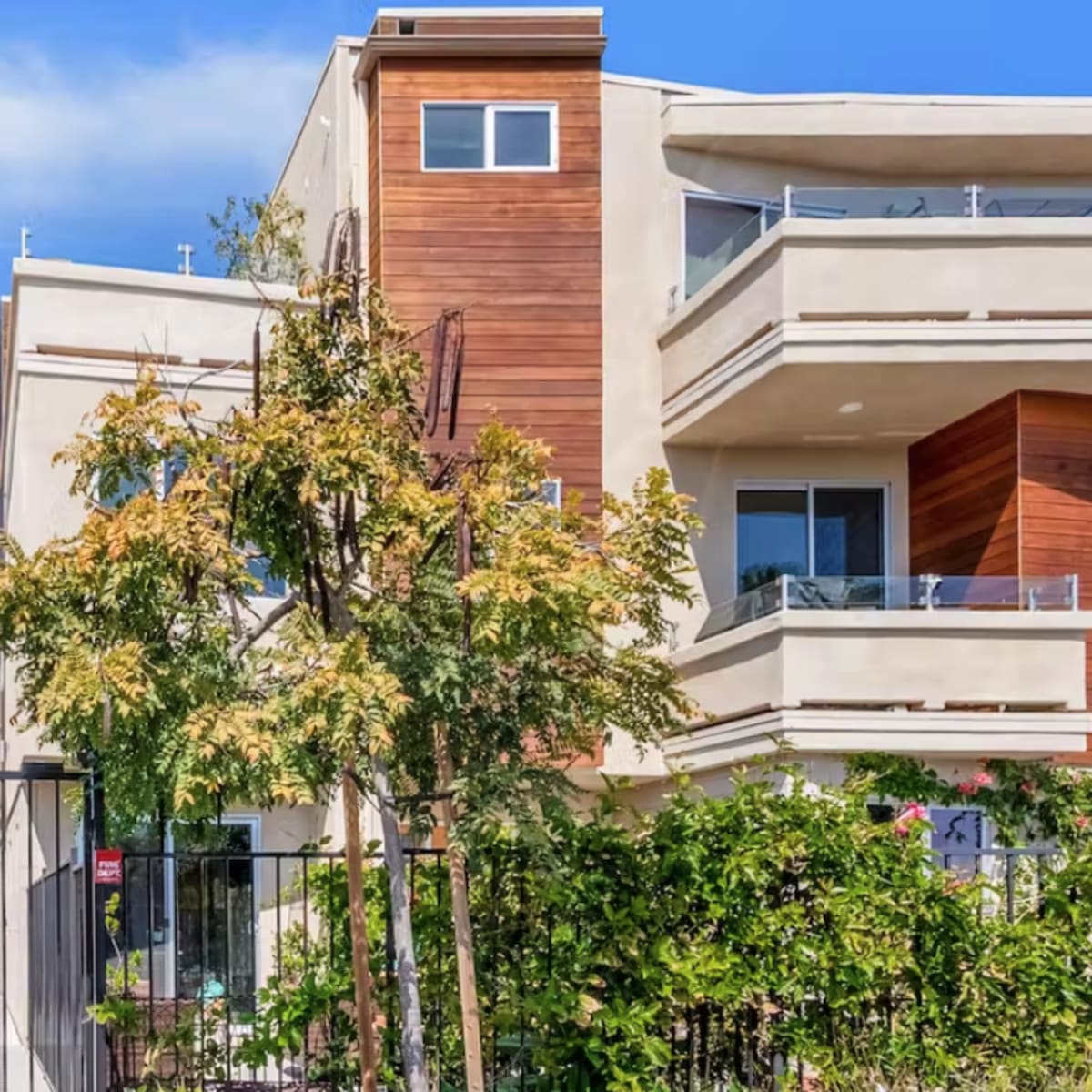 A modern three-story building features a combination of stucco and wooden paneling, with balconies and surrounding greenery.