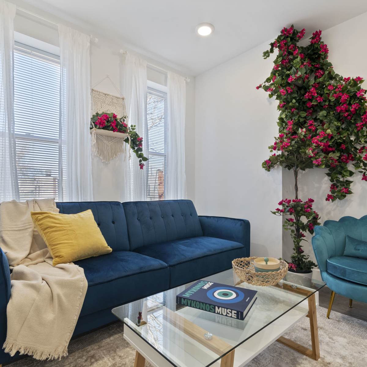 A cozy living room with a blue sofa, a light blue armchair, a glass coffee table, and decorative plants on the wall.