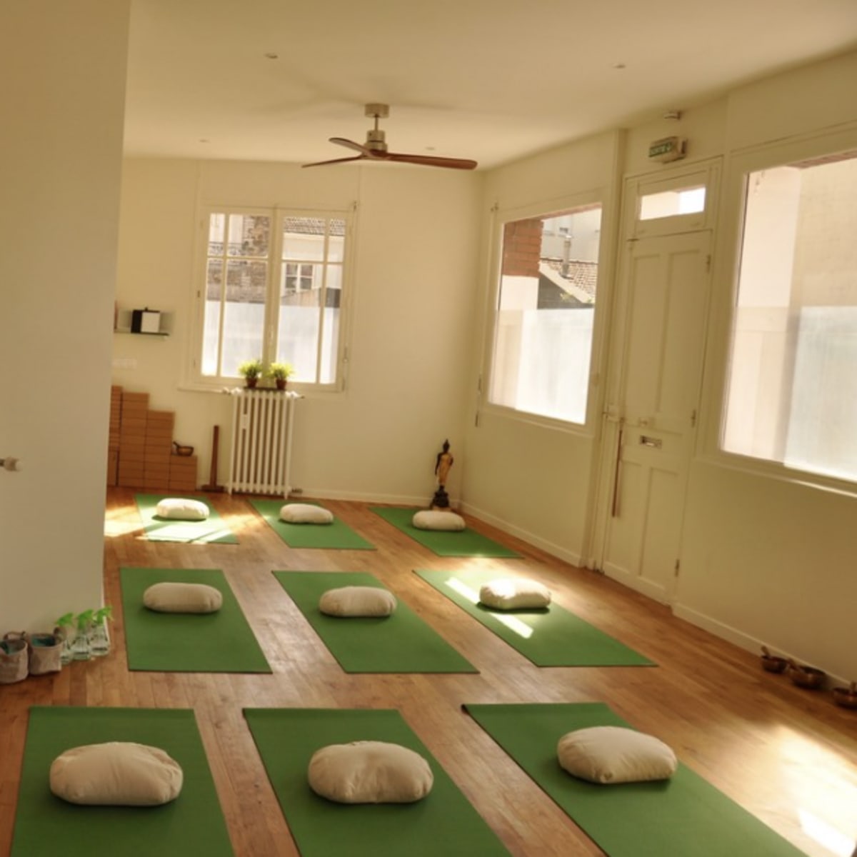 A yoga studio with green mats arranged on a wooden floor, each mat accompanied by a white cushion, and natural light entering through large windows.