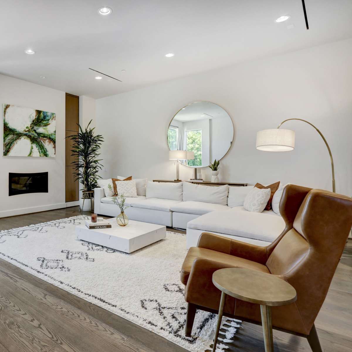 A modern living room features a white sectional sofa, a round mirror, and a wooden coffee table on a patterned rug, complemented by plants and decorative items.