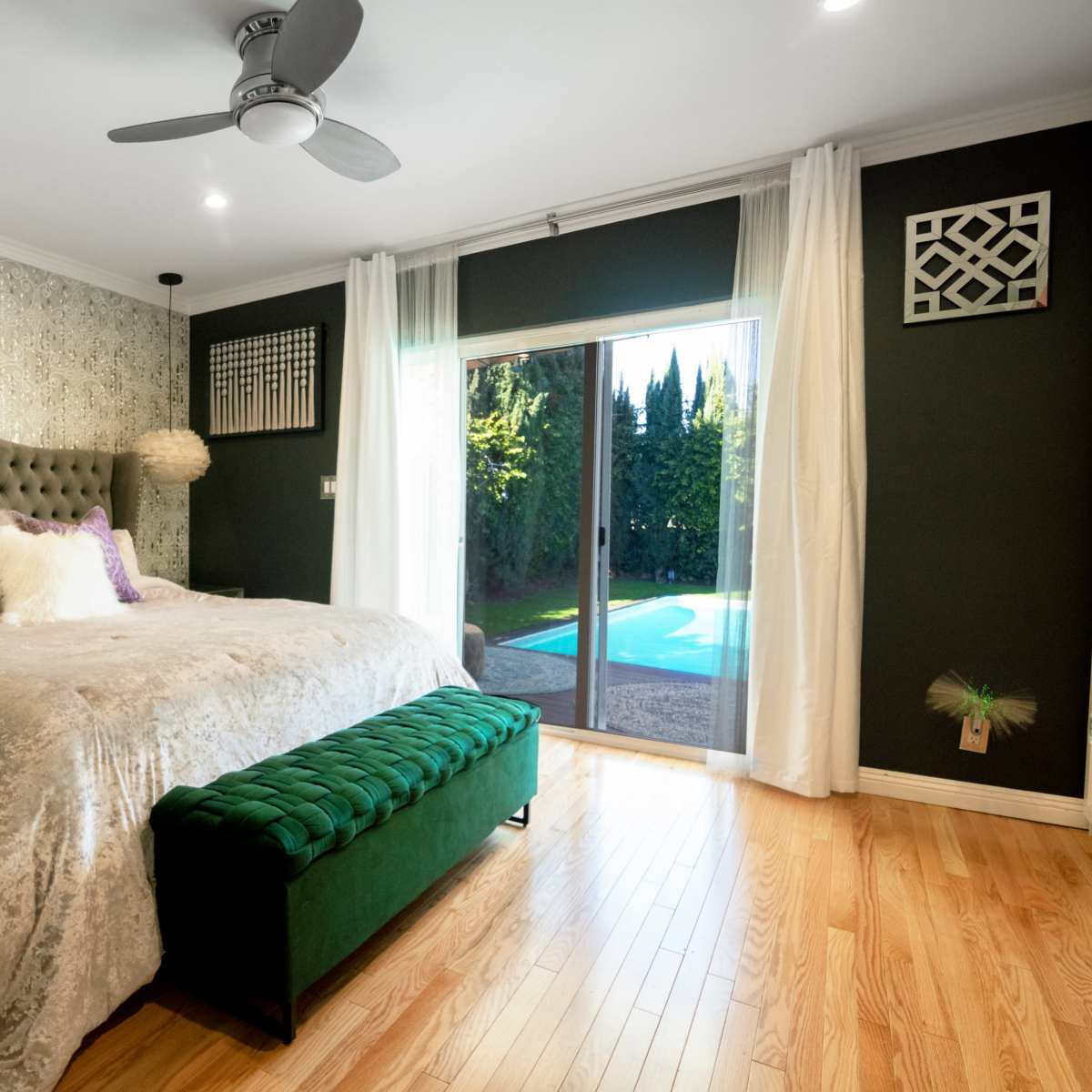 A modern bedroom with a large bed, a green velvet bench, and a sliding glass door that opens to a pool area surrounded by trees.