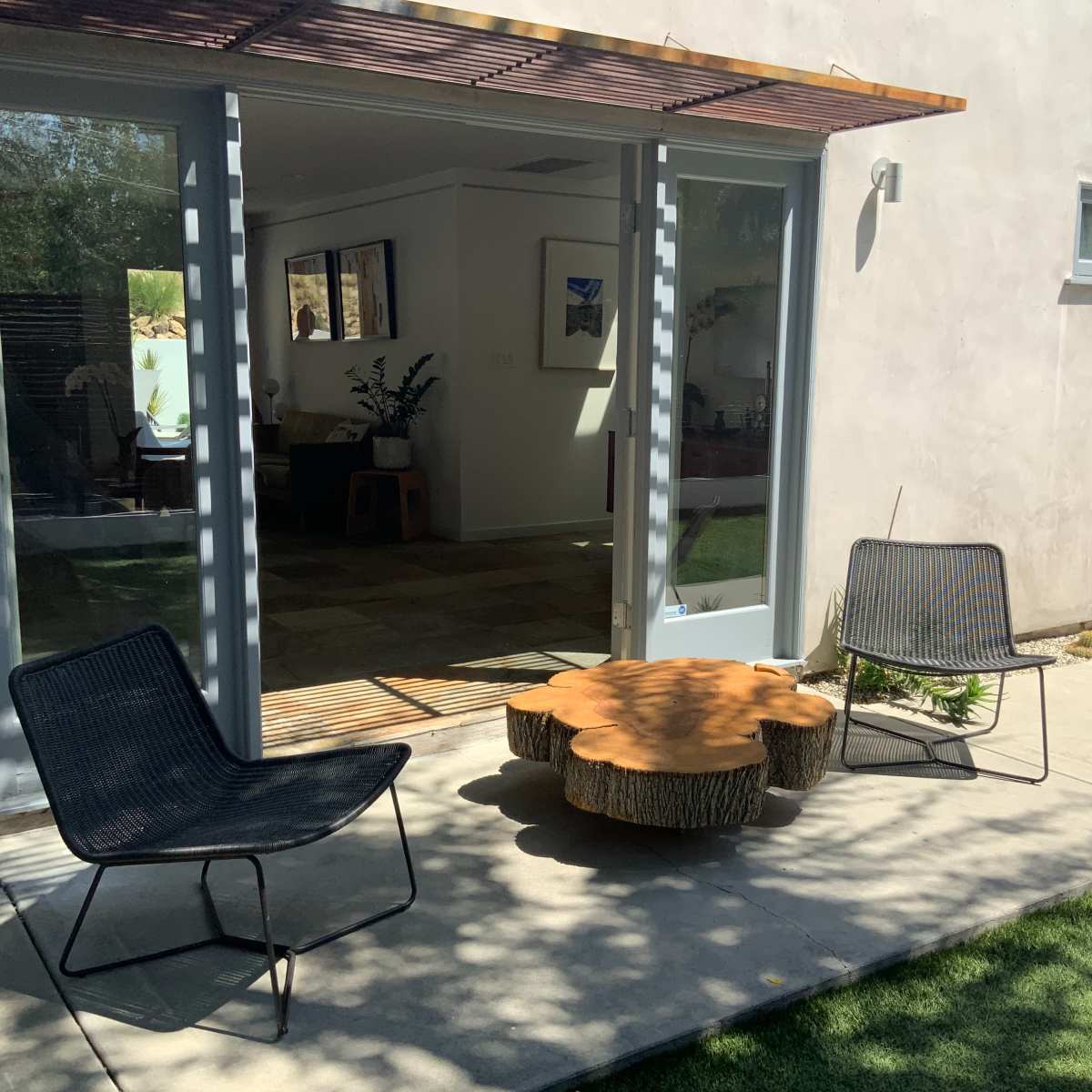 A small outdoor seating area with two black chairs and a round wooden table in front of a house with large glass doors.