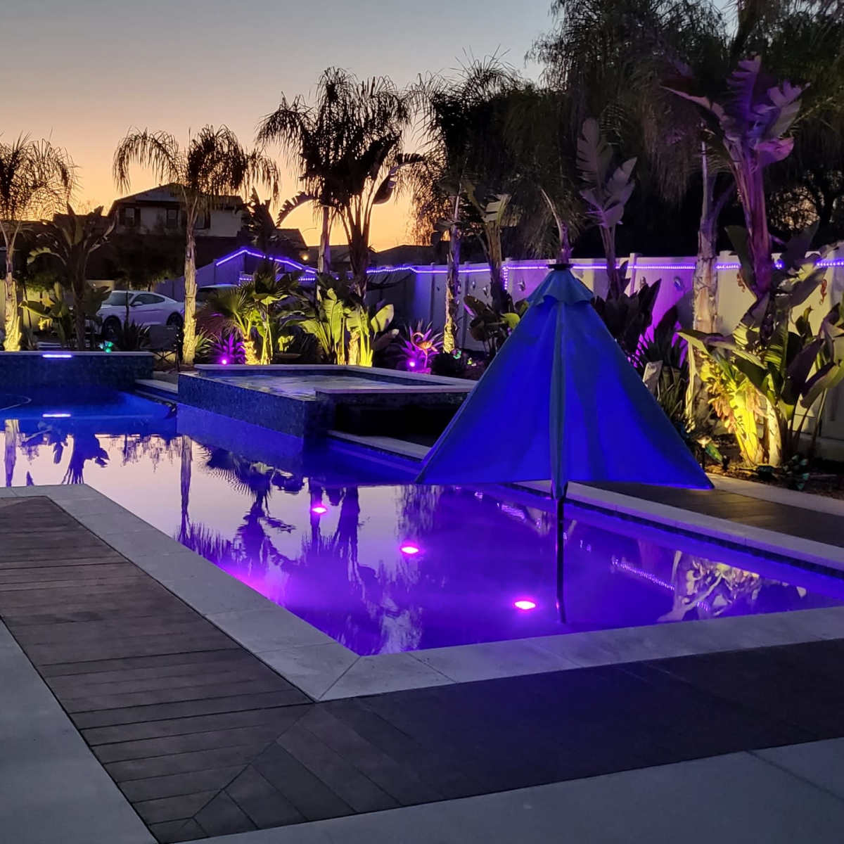 A backyard pool area features illuminated water and plants, with a triangle canopy set up beside the pool at dusk.