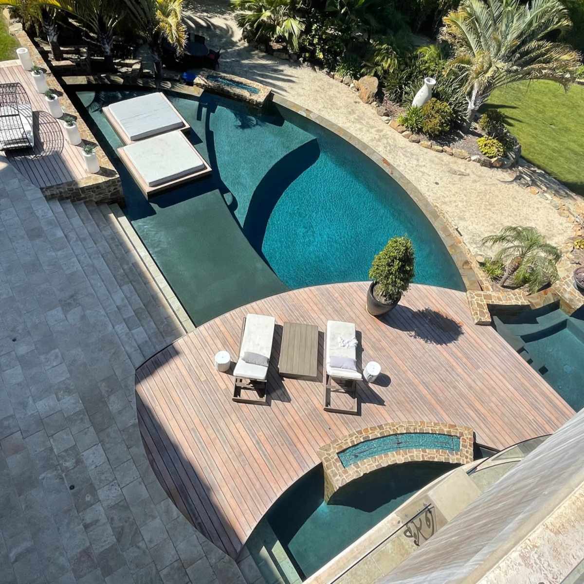 A spacious outdoor area with a curved swimming pool, wooden deck, and lounge chairs surrounded by tropical landscaping.