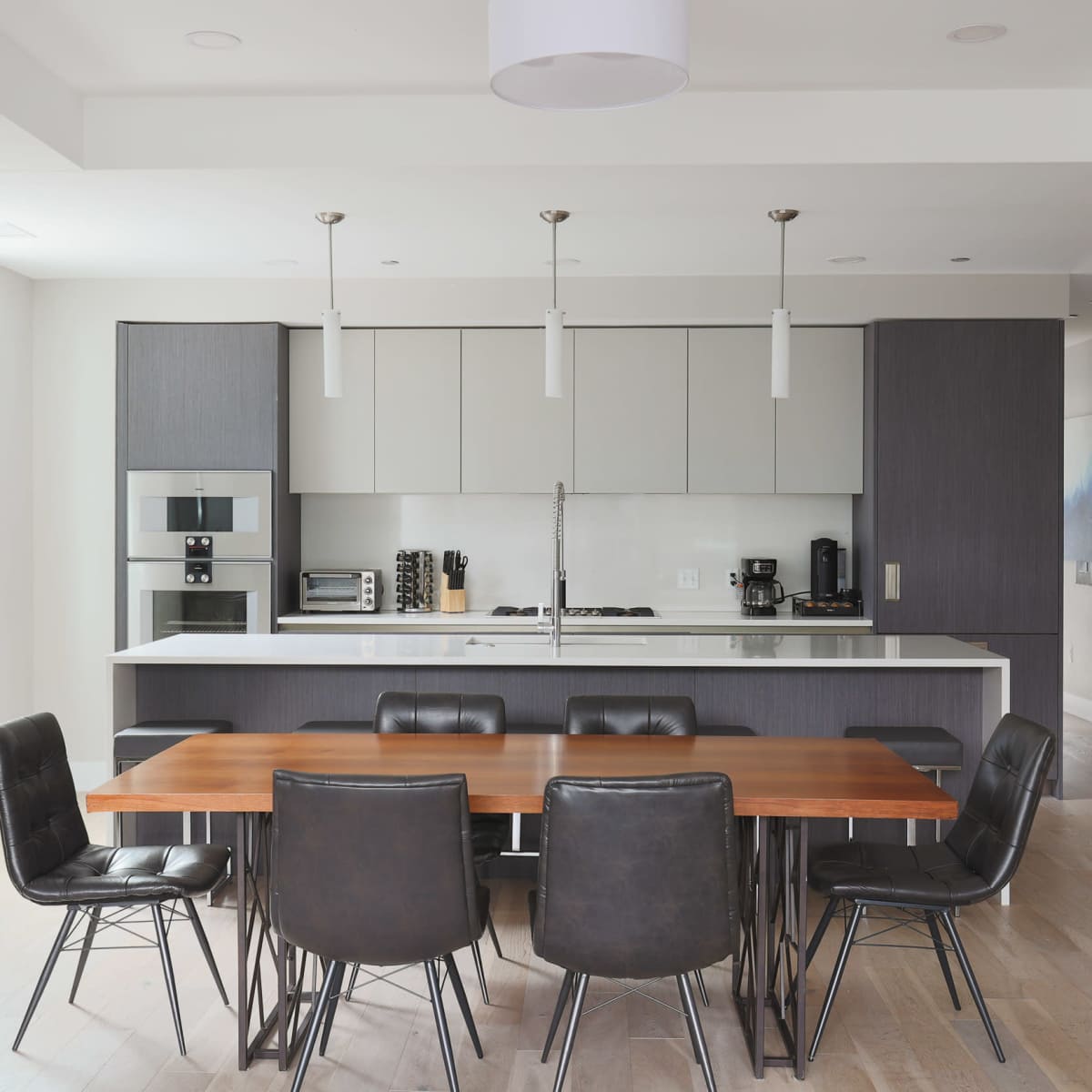 A modern kitchen and dining area features a long wooden table surrounded by black leather chairs, with a sleek kitchen cabinet and appliances in the background.