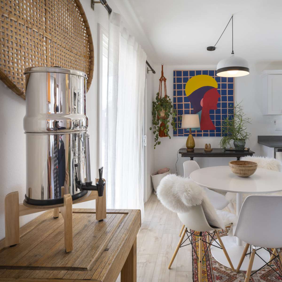 A modern kitchen and dining area features a round table with white chairs, a stainless steel water filter, and a colorful wall art piece.