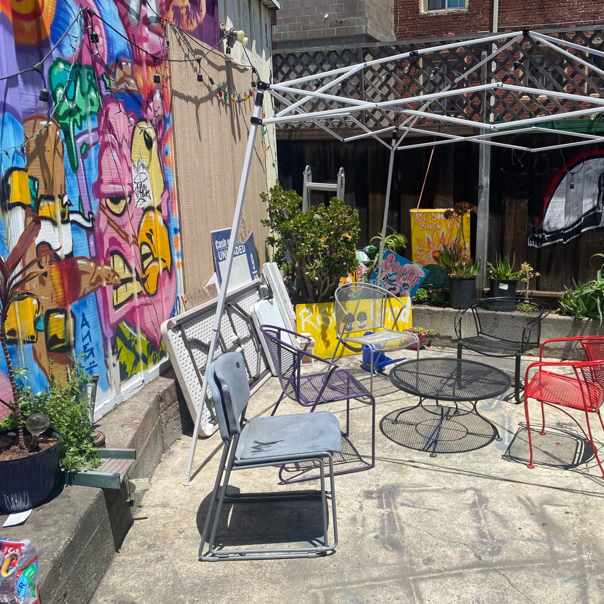 A shaded seating area with various chairs and tables is set up beside a colorful mural and potted plants.