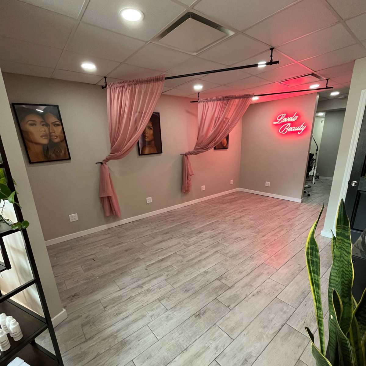 A beauty salon interior with light-colored wooden flooring, pink curtains, and a neon sign reading "Dani's Beauty" on the wall, alongside framed portraits of two women.