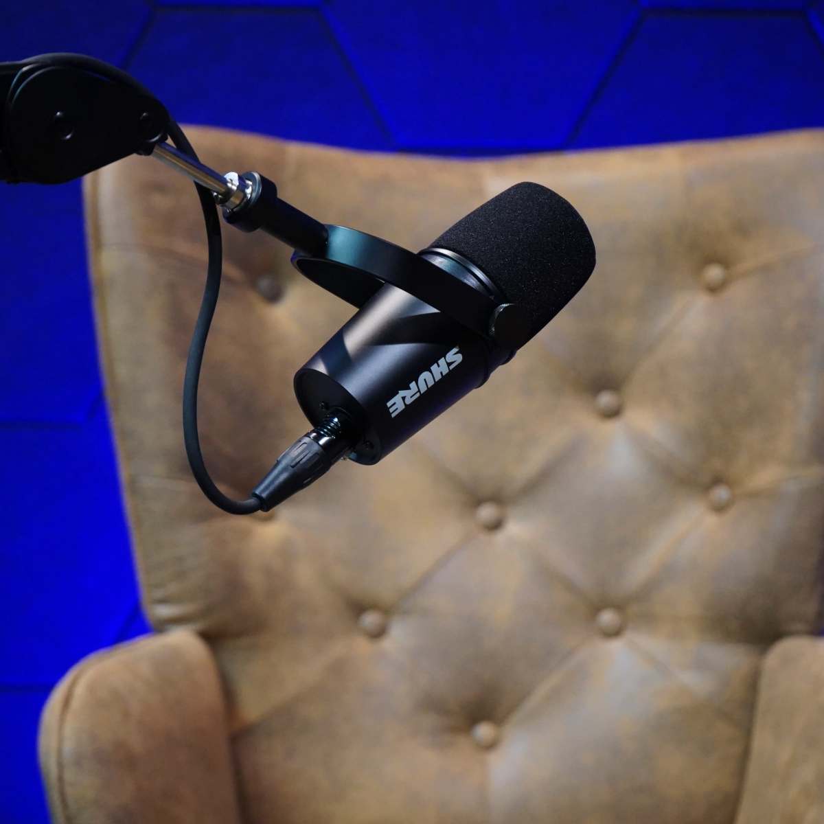 A microphone is mounted on a black arm above a brown leather chair in front of a blue, hexagonal-patterned backdrop.