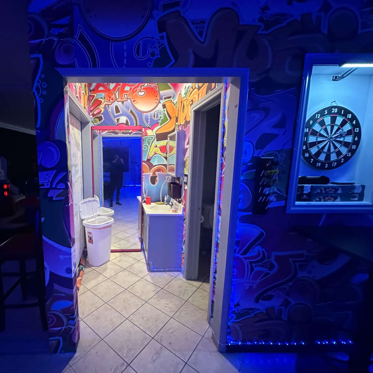 A vibrant hallway with colorful graffiti on the walls, leading to a room with a dartboard and a bin visible in the kitchen area.