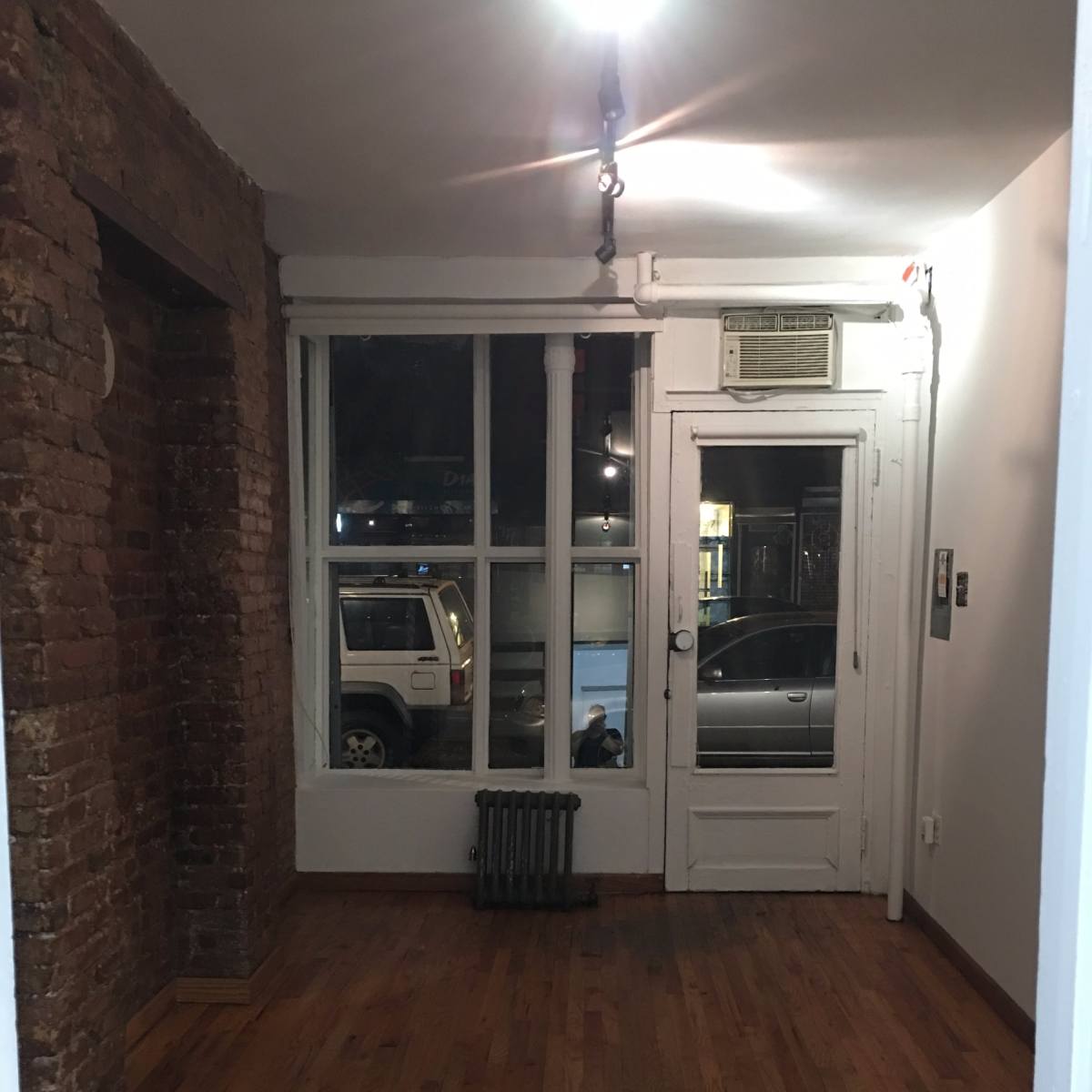 A sparsely furnished room with exposed brick walls, a radiator, a window revealing a street view, and a door leading outside.