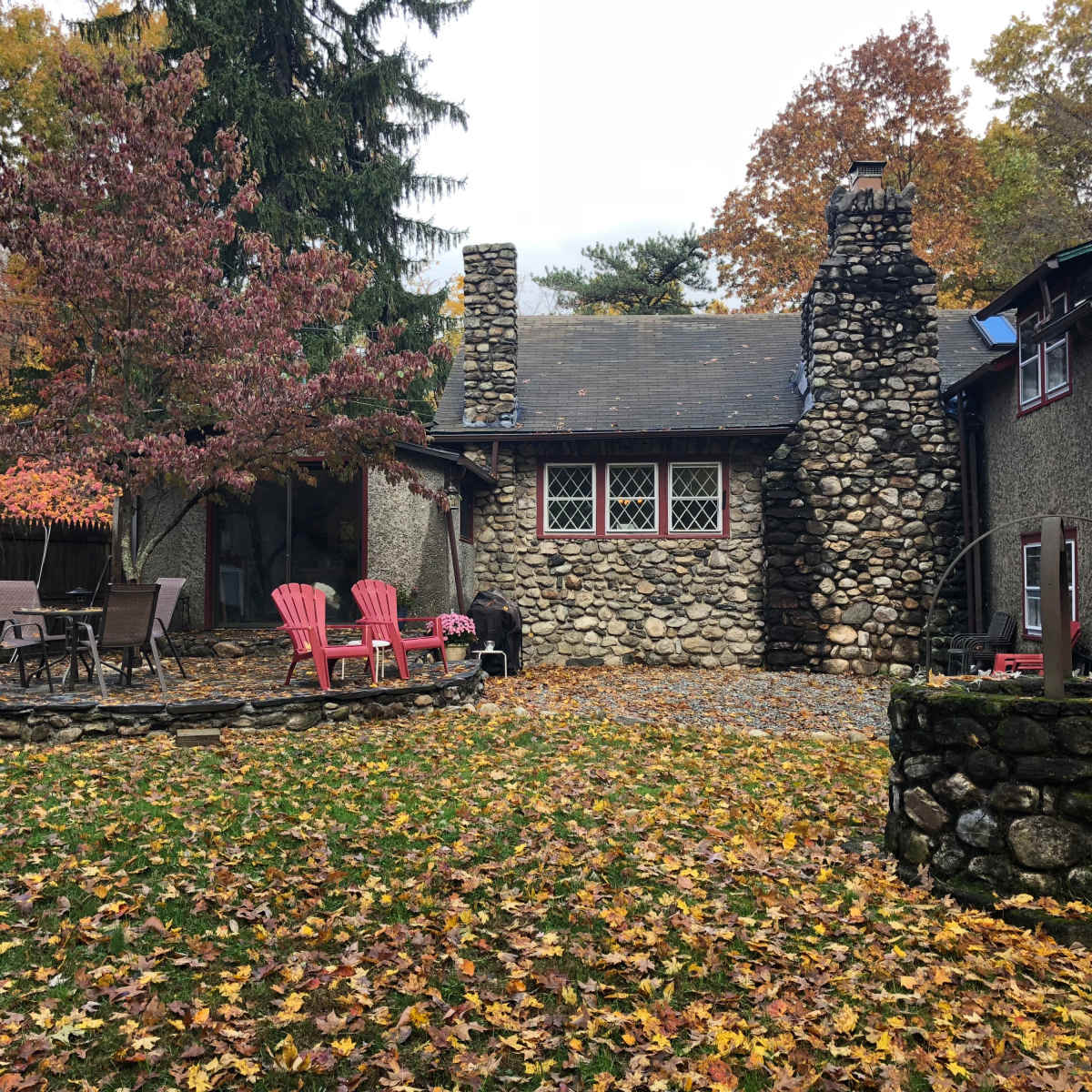 A stone cottage surrounded by trees and a yard covered in fallen leaves, featuring pink chairs and a circular stone fire pit.