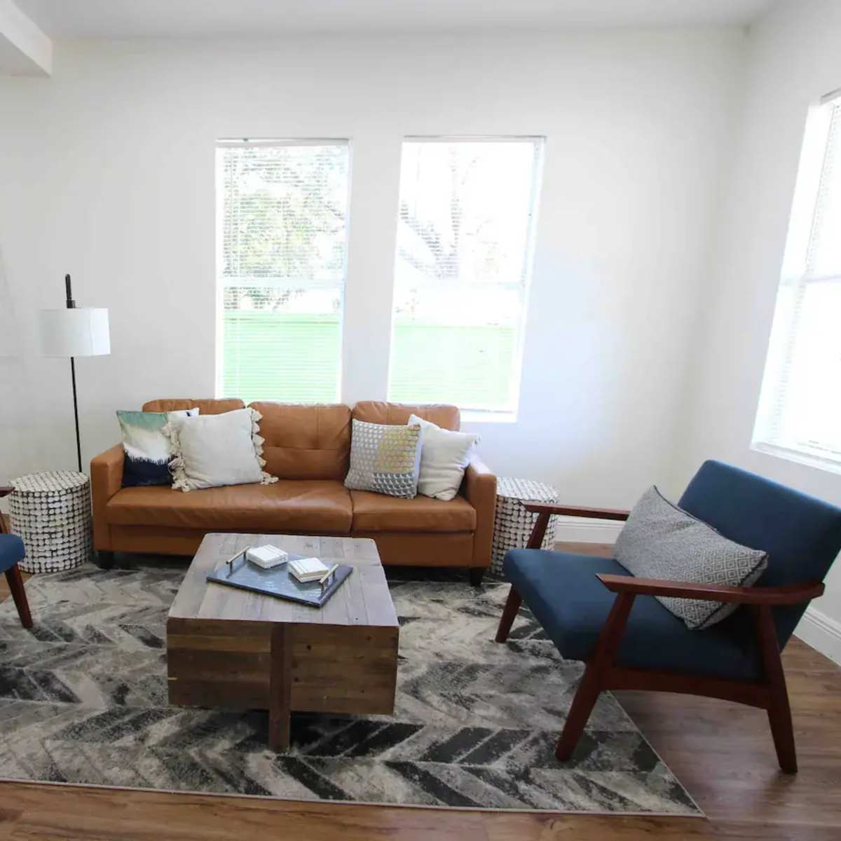 The scene features a modern living room with a brown leather sofa, two blue armchairs, a wooden coffee table, and large windows allowing natural light to enter.