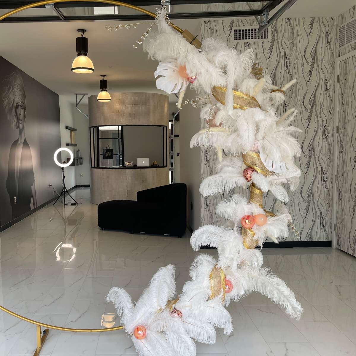A modern indoor space featuring a decorative gold circular frame adorned with white feathers and floral accents, alongside a reception area with a black sofa and lighting equipment.