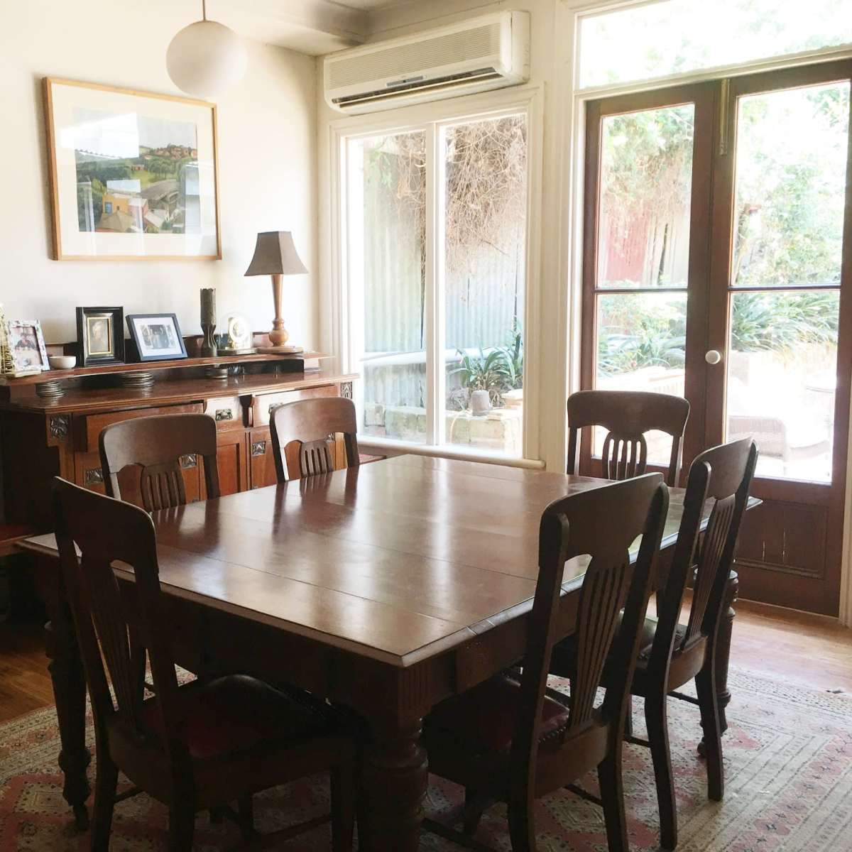 A dining room with a wooden table surrounded by eight chairs, a sideboard with framed photos, and large windows leading to an outdoor area.