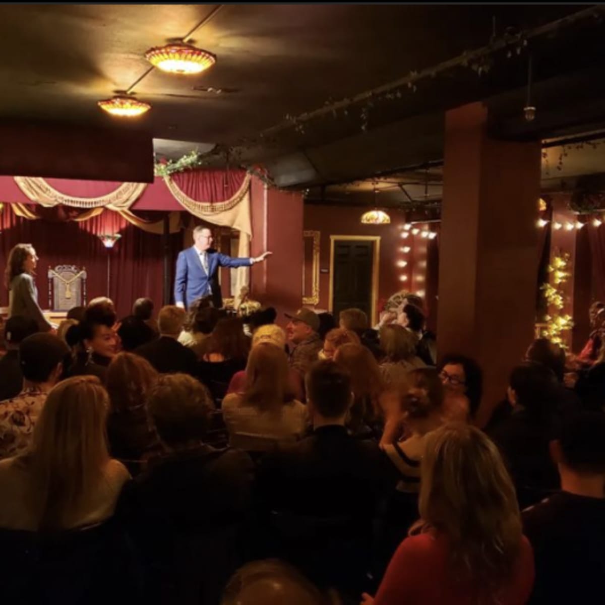 A crowded room with a stage featuring a performer speaking to an audience amidst dim lighting and decorative drapery.