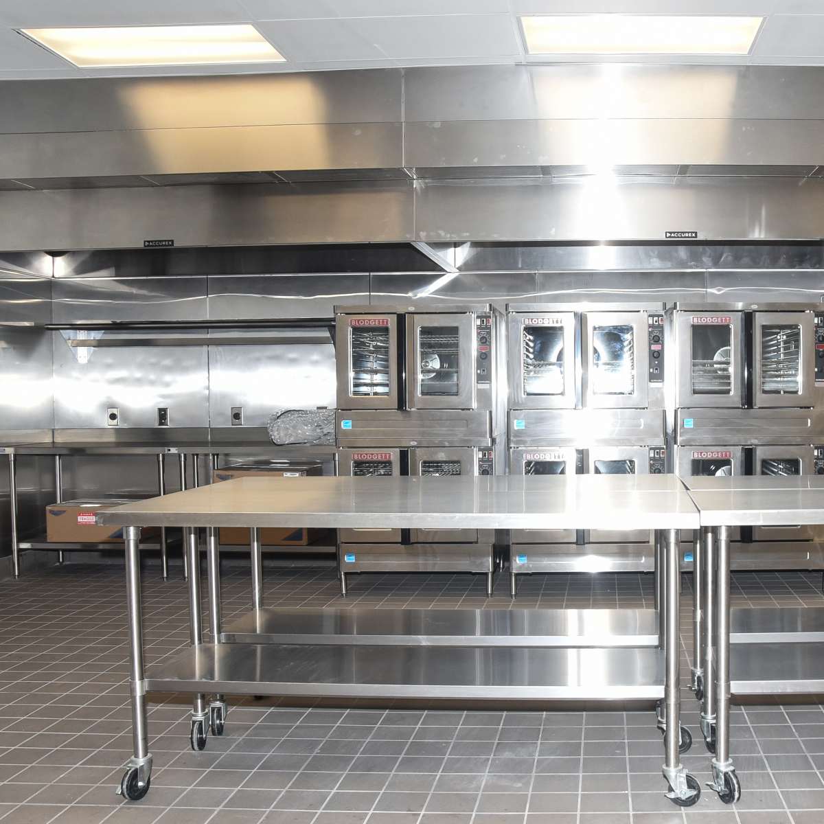 A professional kitchen with stainless steel appliances and countertops, featuring several ovens and open shelving.
