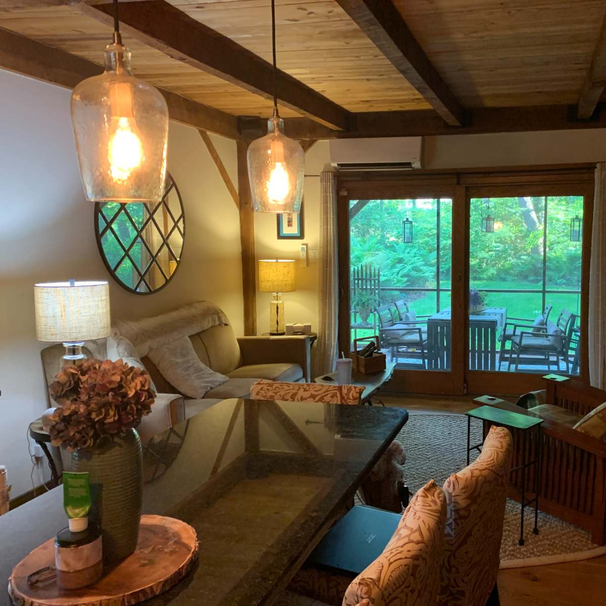 A cozy living space featuring wooden beams, a dark dining table, comfortable seating, and large windows overlooking a green outdoor area.