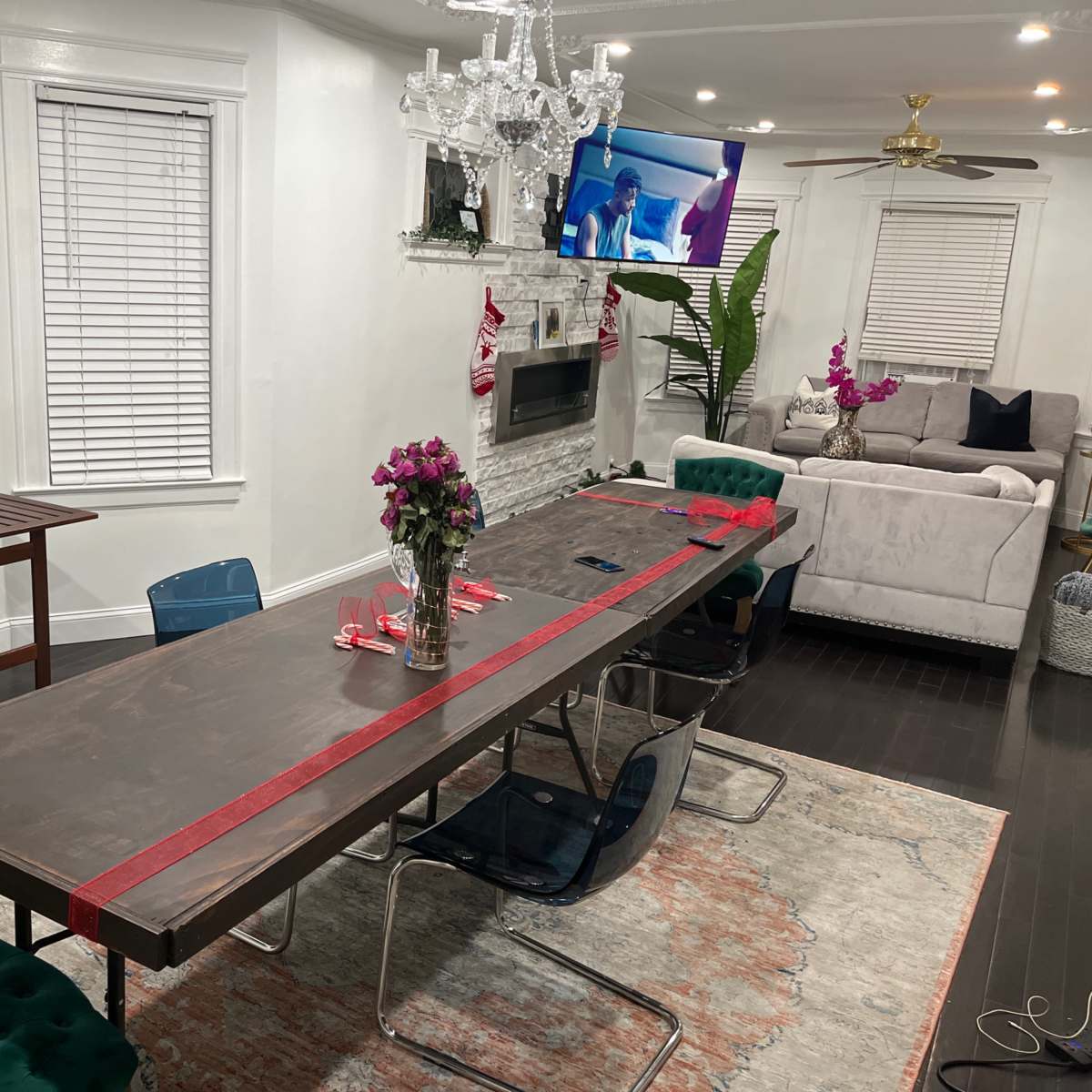 A long dining table is set with red ribbon and flowers, surrounded by chairs, in a room featuring a television, a sofa, and decorative plants.