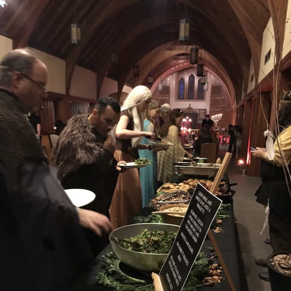 A group of individuals dressed in costumes serves themselves from a long buffet table filled with food in a dimly lit hall with wooden arches.