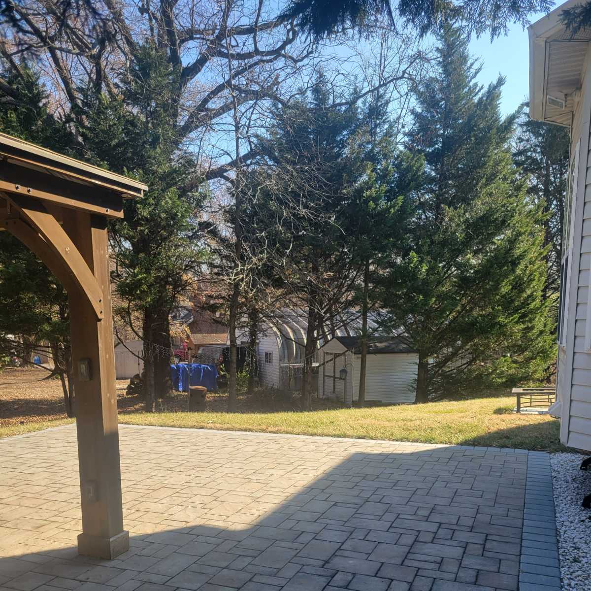 A paved backyard area next to a wooden structure, surrounded by trees and a grassy patch, with a shed visible in the background.