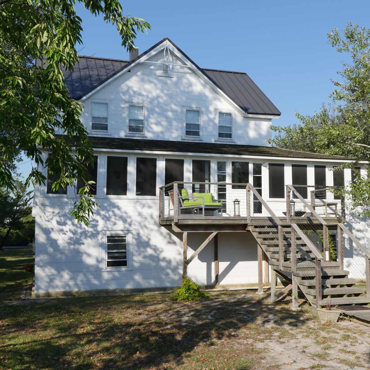 A two-story white house with a dark metal roof features a front porch and wooden stairs leading to a deck.