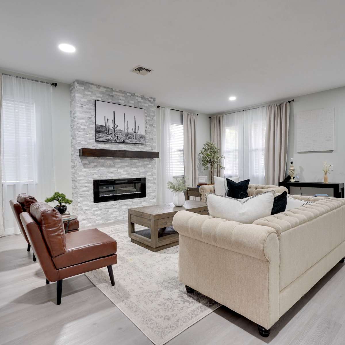 The living room features a cozy arrangement with a beige sofa, a wooden coffee table, and two brown leather chairs, set against a stone fireplace and large windows with sheer curtains.