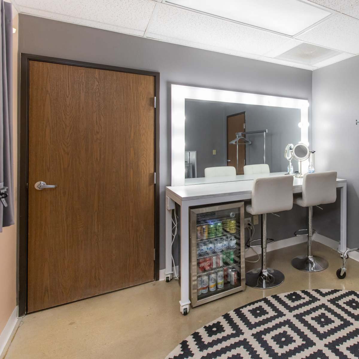 A modern dressing room featuring a makeup vanity with illuminated mirrors, a rack for clothes, and a mini fridge on a patterned rug.