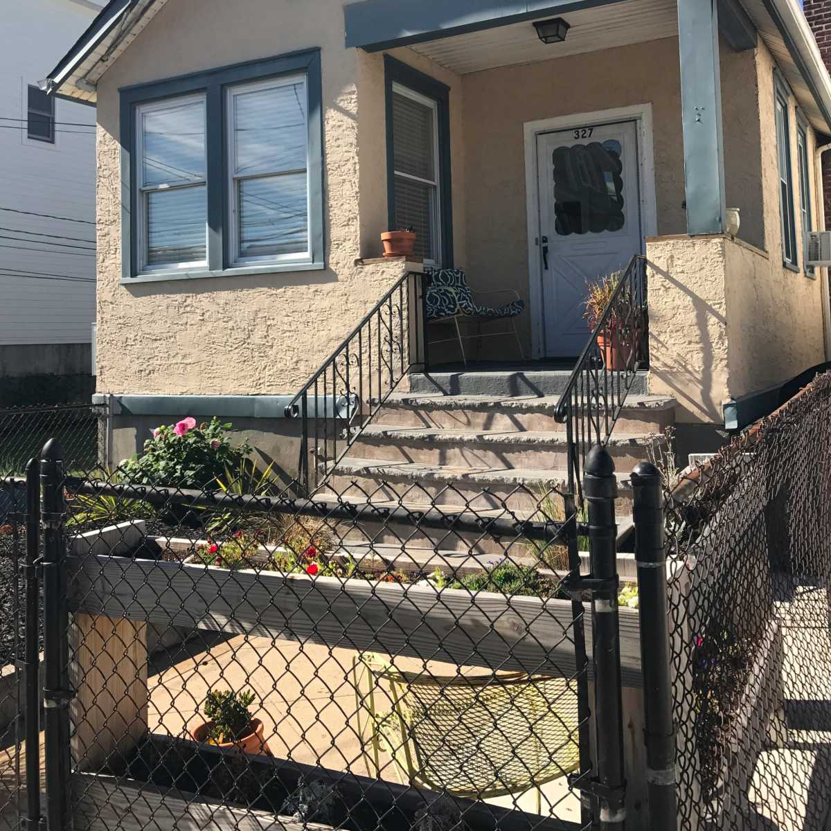 A small, single-story house with a front porch, surrounded by a fence and featuring a garden area.
