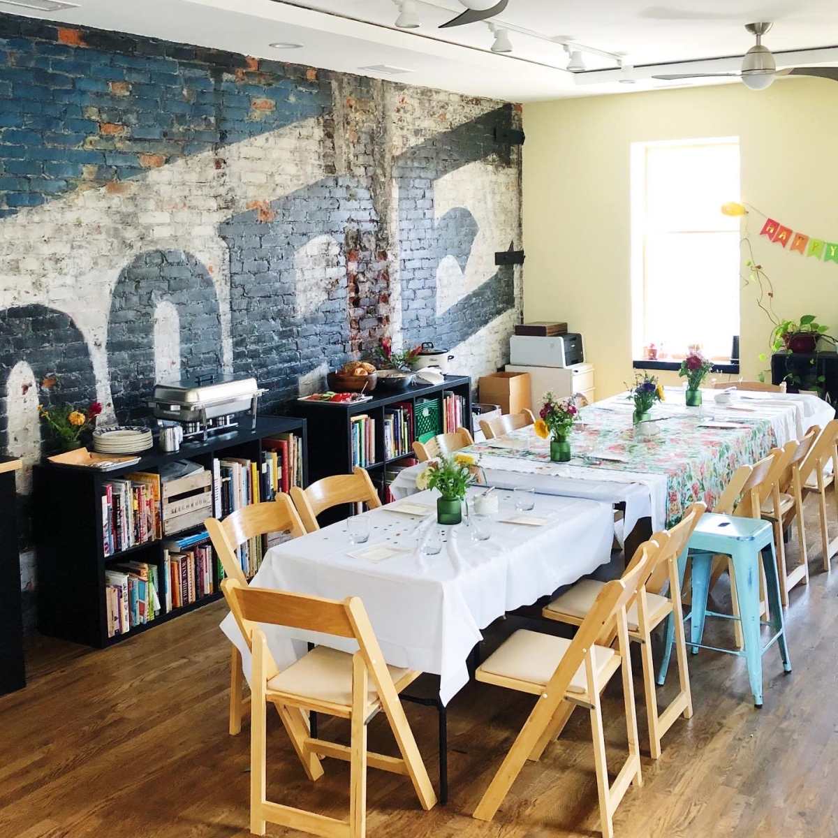 A dining area features multiple tables set with flowers, surrounded by bookshelves and a brick wall displaying a faded mural.