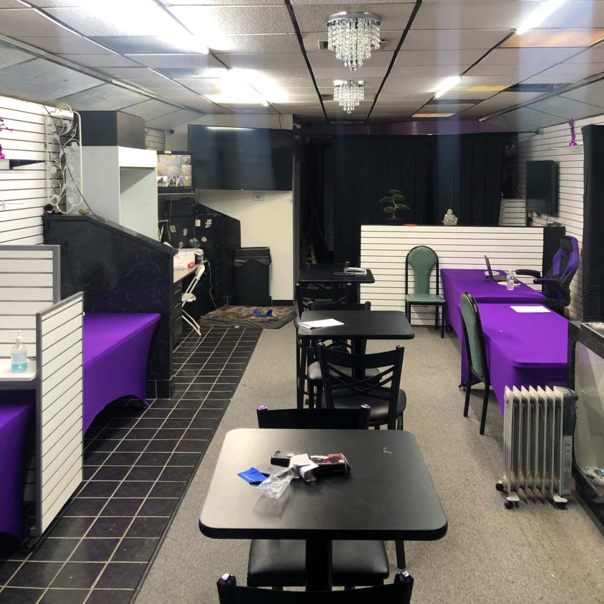 The interior of a small cafe or event space featuring empty tables with purple table covers, chandeliers, and a black curtain at the back.