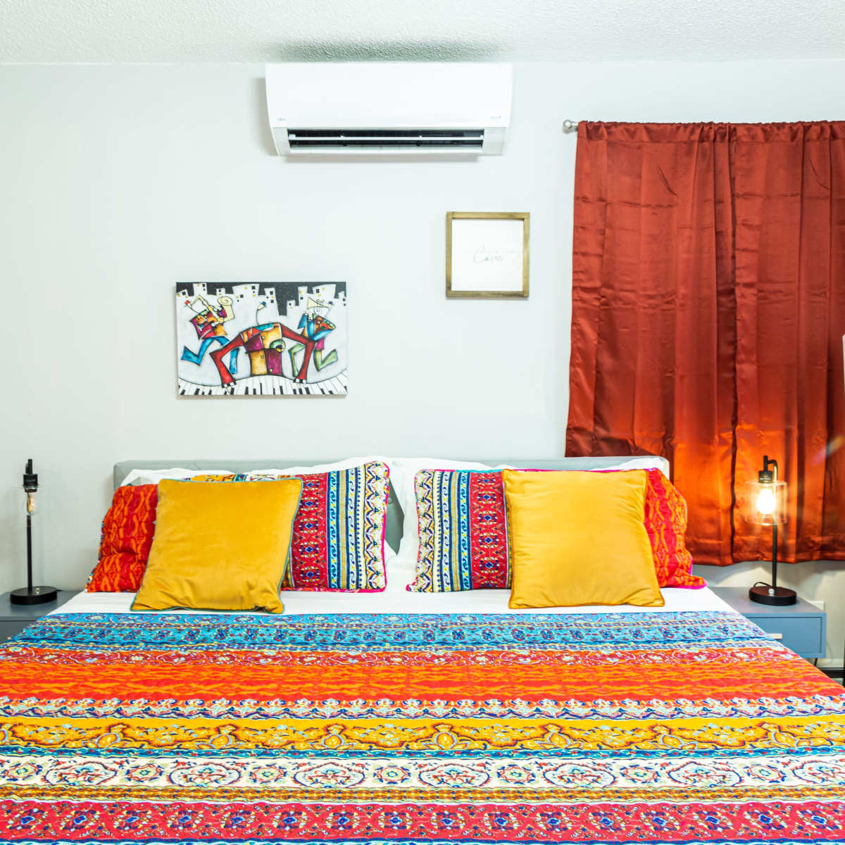A colorful bedspread with decorative pillows is showcased in a well-lit bedroom featuring artwork and curtains.