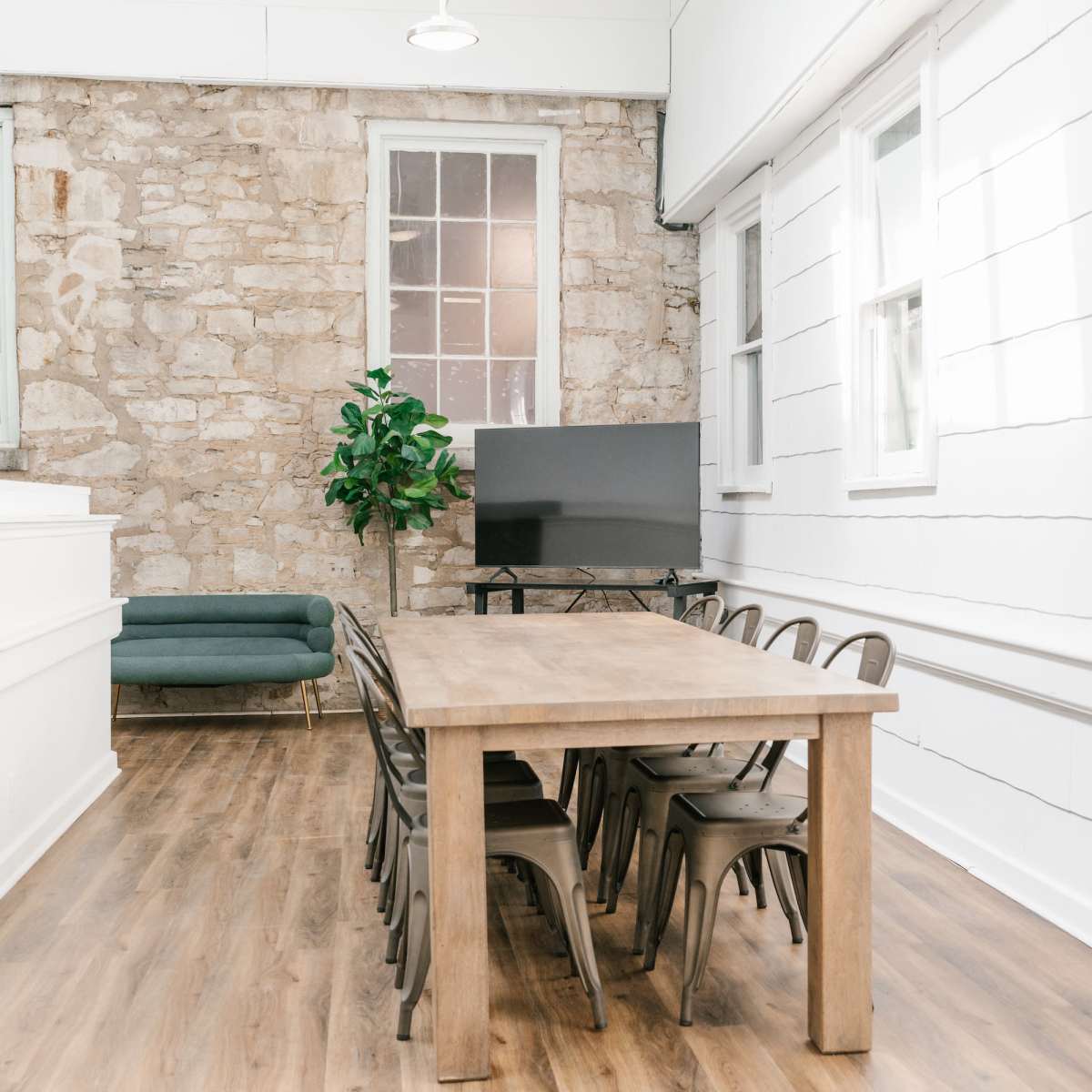 A spacious room featuring a wooden dining table surrounded by metal chairs, with a green sofa and a television against a stone wall.