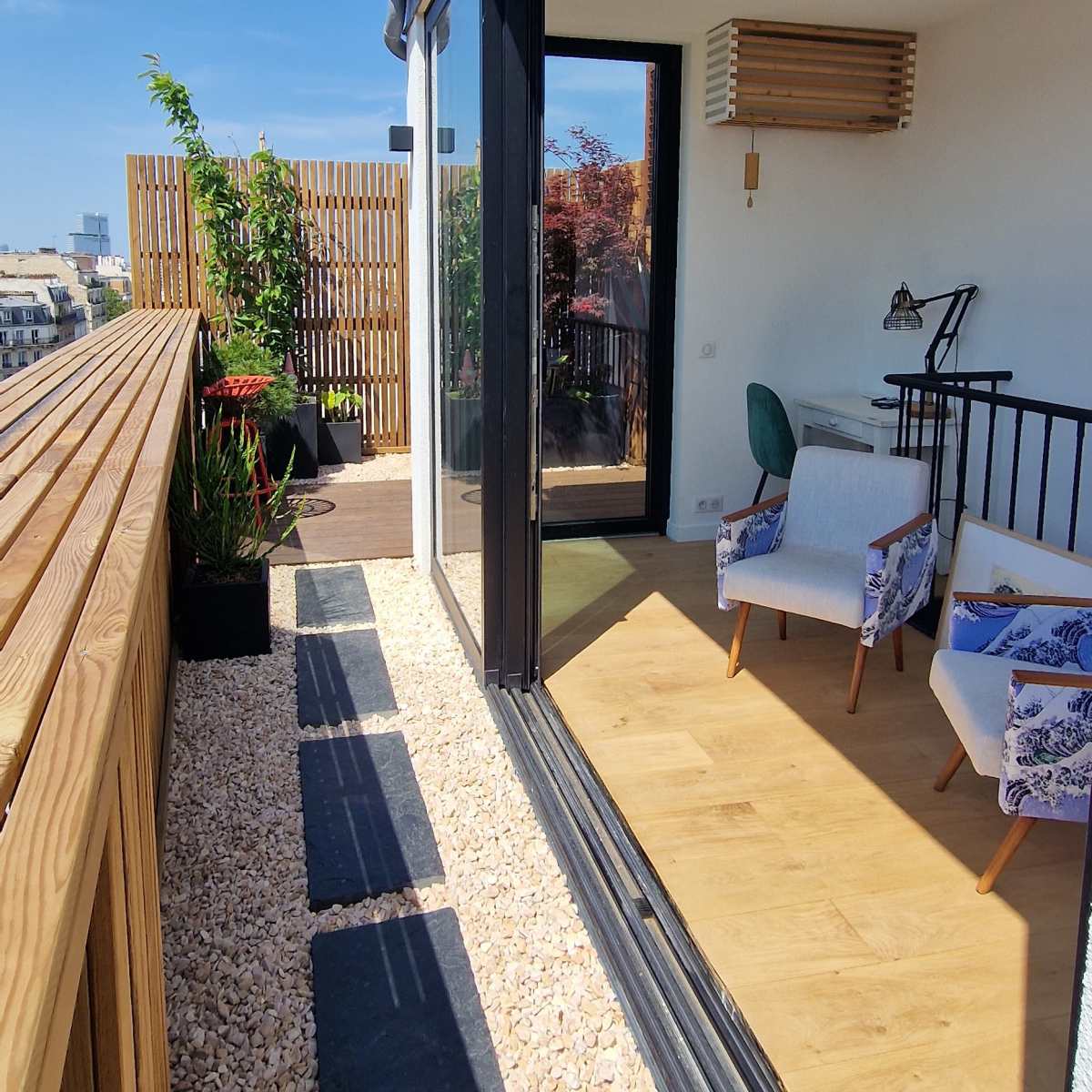 A modern balcony with a wooden railing and paved walkway leads to an indoor space featuring a chair and desk, with clear skies in the background.