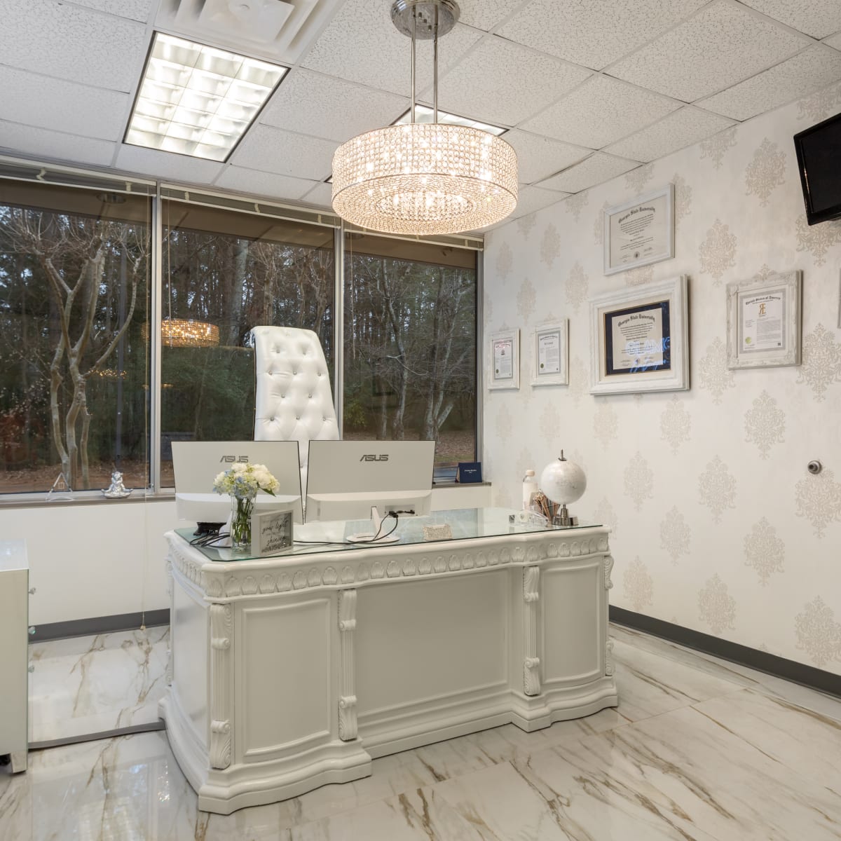 A modern office interior with a white desk, a high-backed chair, and a decorative chandelier, surrounded by framed certificates and a large window.