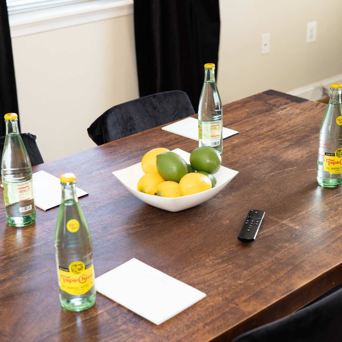 A wooden table is set with sparkling water bottles, coasters, and a bowl of lemons and limes at the center.