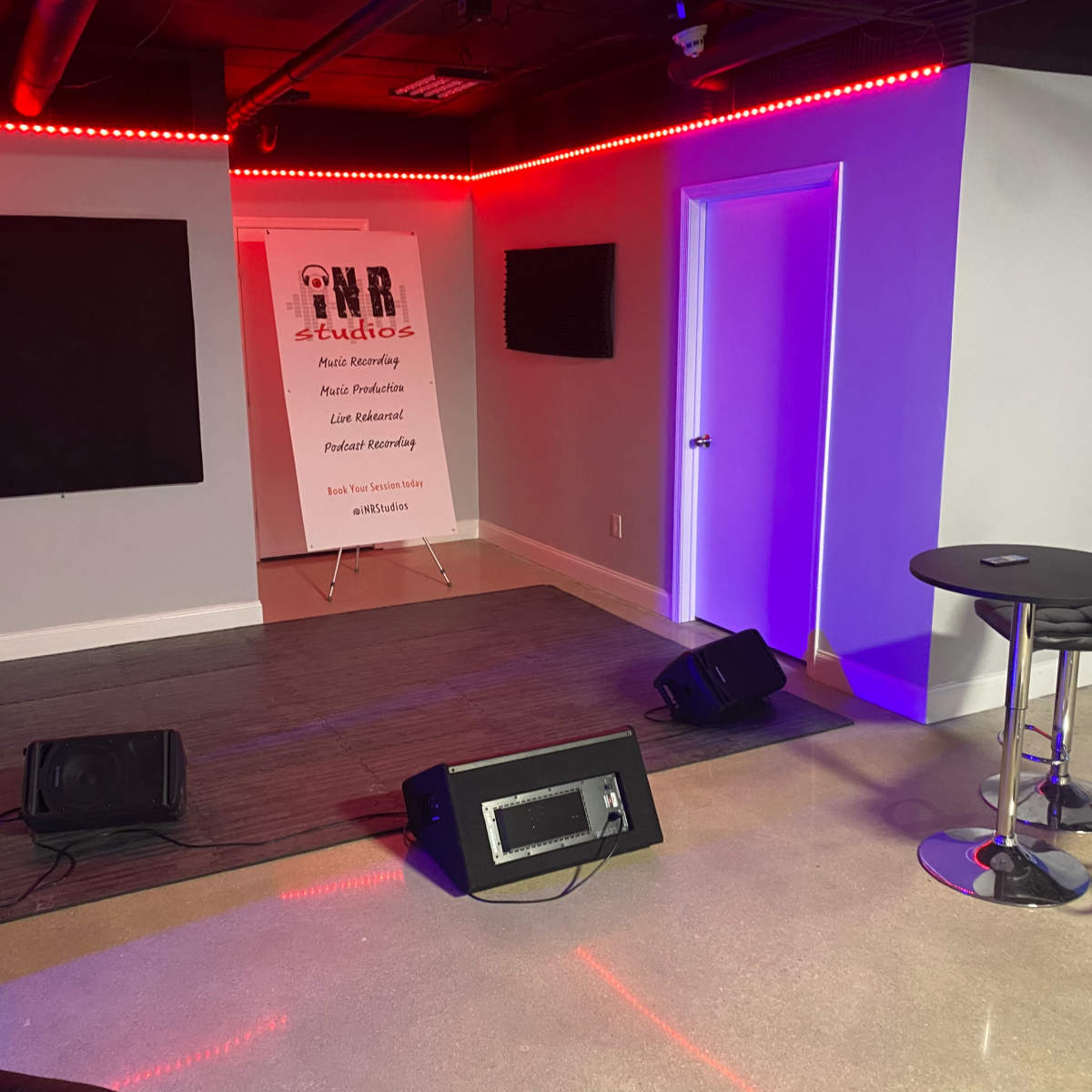 A small music studio with a stage area, featuring a microphone stand, sound equipment, and a backdrop with text, surrounded by colorful lighting and seating.