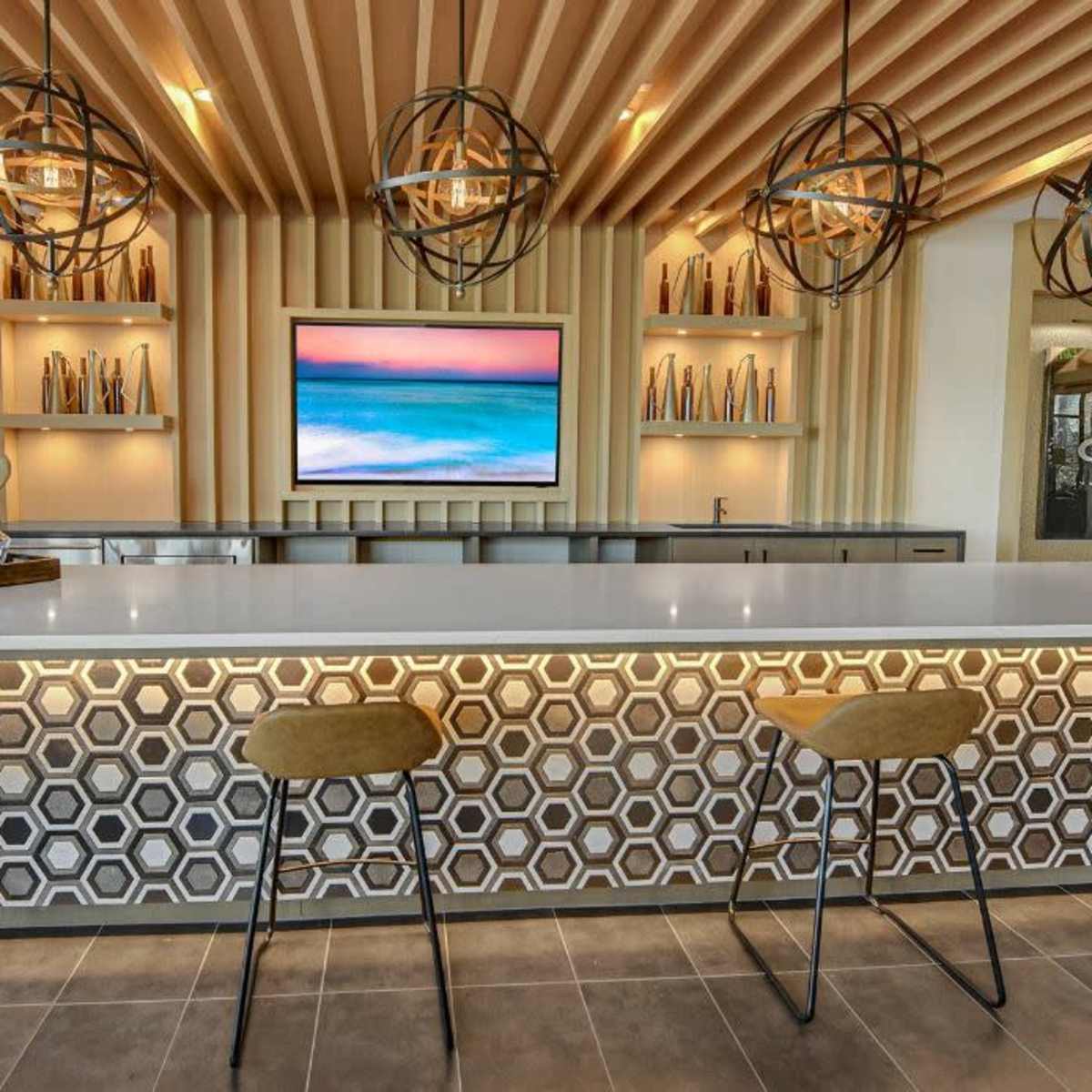 A modern bar area with a white countertop, geometric patterned front, and pendant lighting above, complemented by a large screen displaying a scenic view.