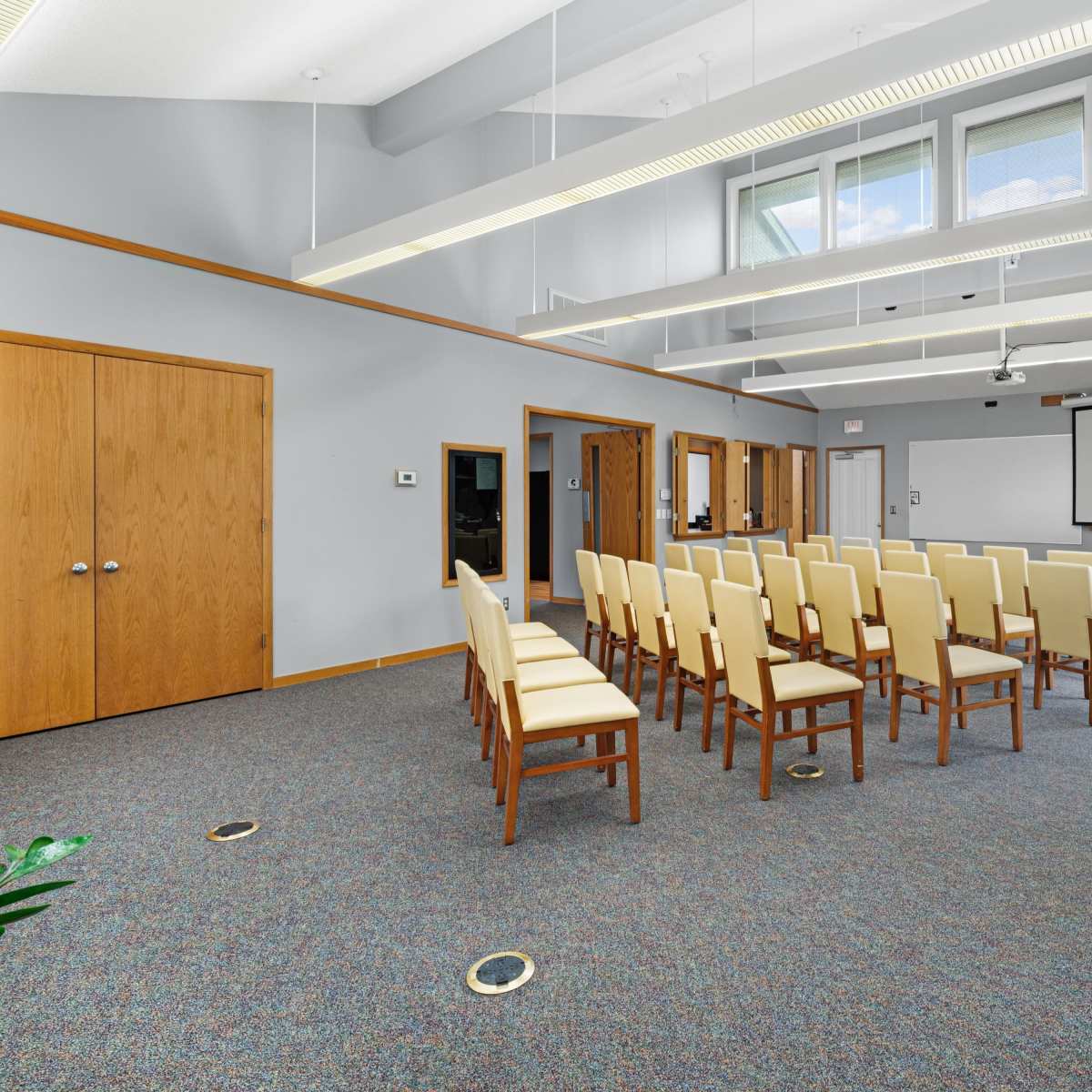 A meeting room features rows of beige chairs arranged neatly in front of a presentation screen, with light-colored wooden doors and large windows along the walls.