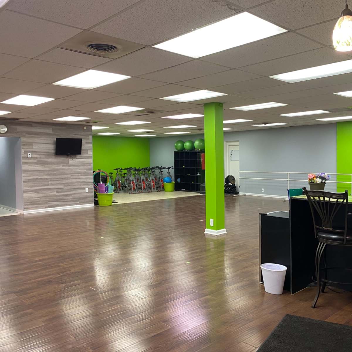 An empty indoor fitness studio with light wood flooring, green accent walls, and fitness equipment visible in the background.