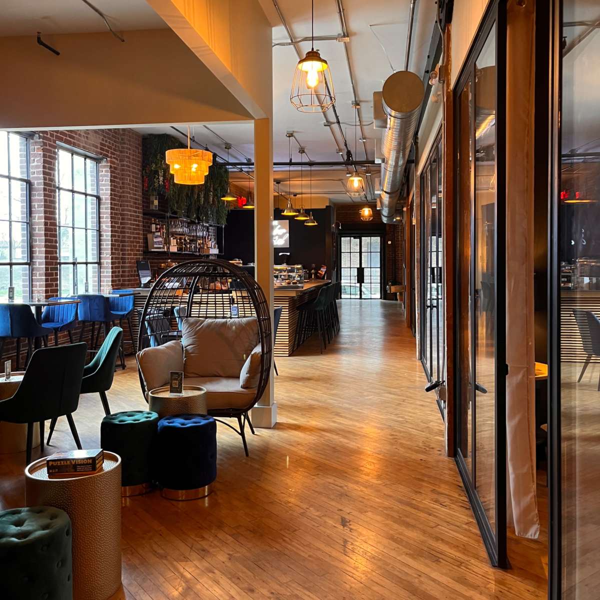 A stylish interior of a modern café or lounge with wooden flooring, industrial-style lighting, and a seating area featuring a hanging chair and colorful stools.