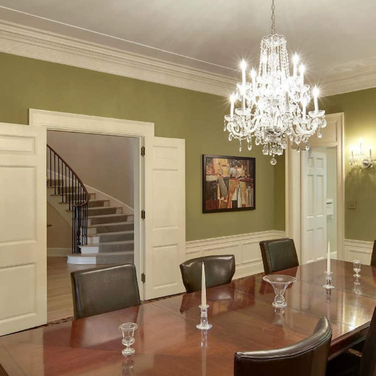 A spacious dining room with a large wooden table, surrounded by chairs, and featuring a chandelier and a staircase in the background.