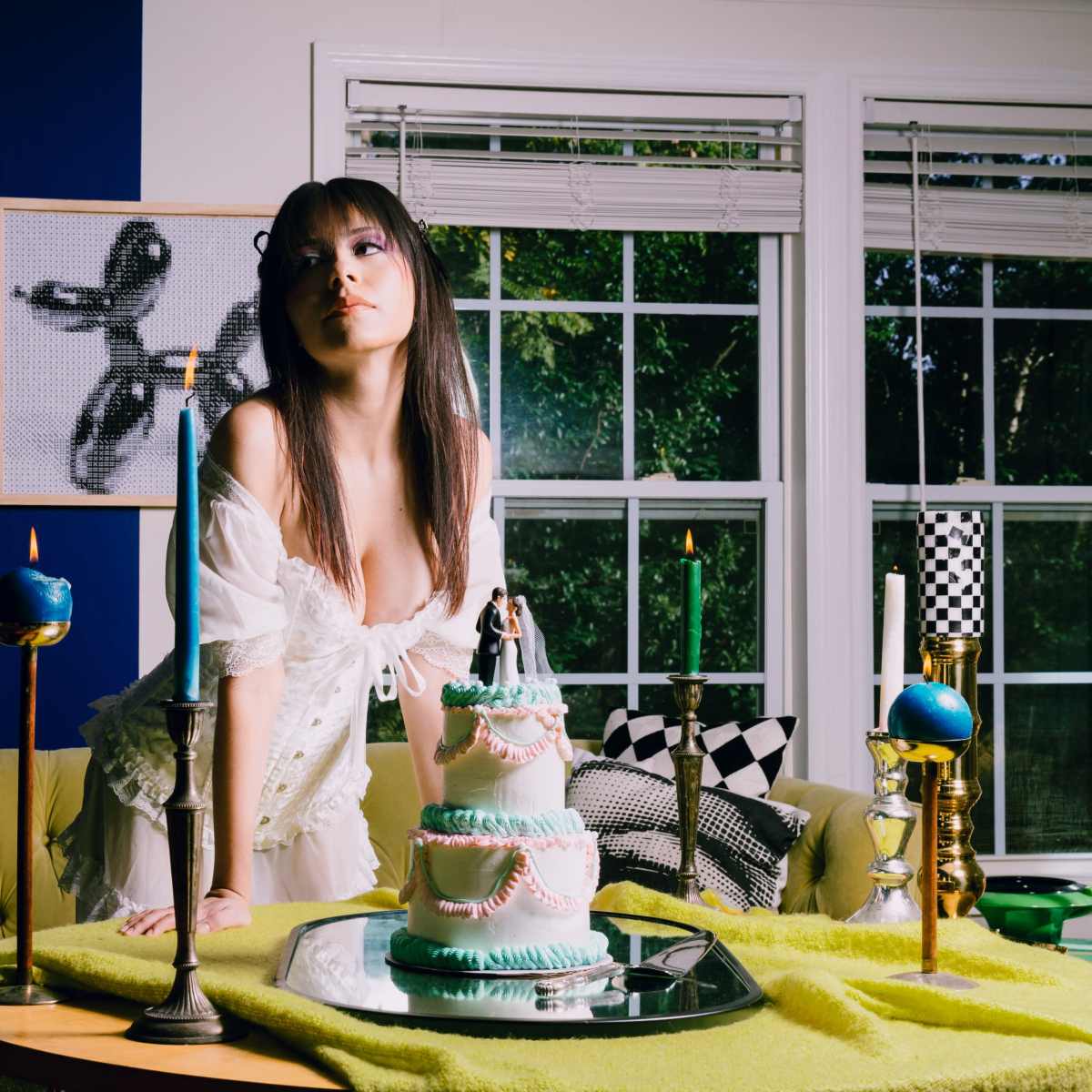 A person stands beside a three-tiered cake on a table adorned with candles and colorful decor in a well-lit room with large windows.