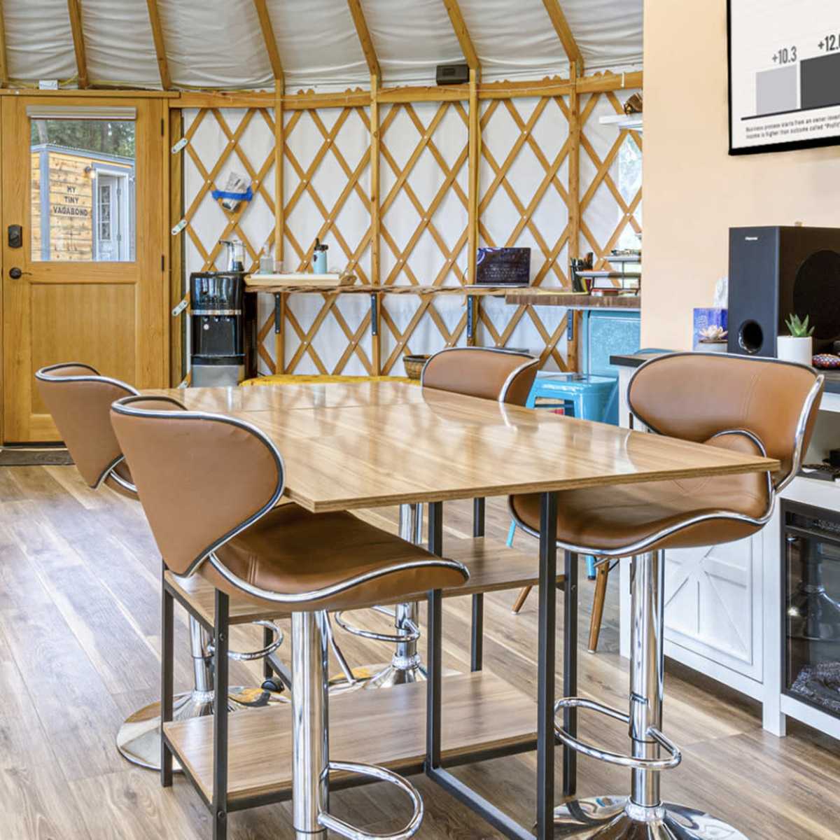 A modern interior space with a wooden table and brown chairs, featuring a wall-mounted screen, a sound system, and a door leading outside.