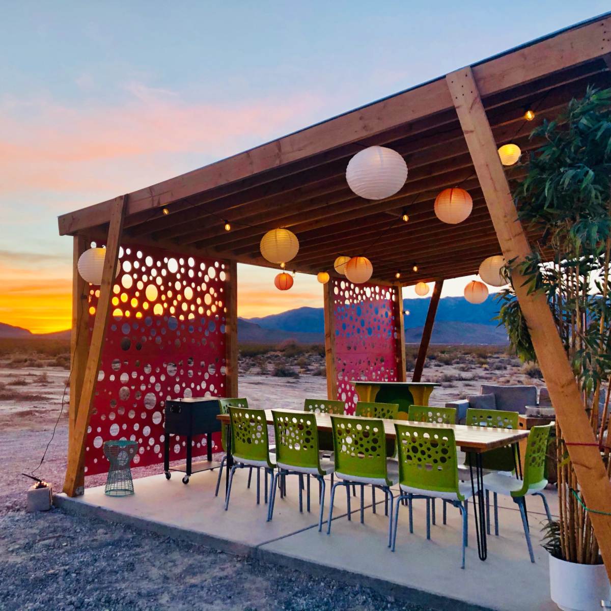 A covered outdoor seating area with colorful chairs and hanging lanterns overlooks a desert landscape at sunset.