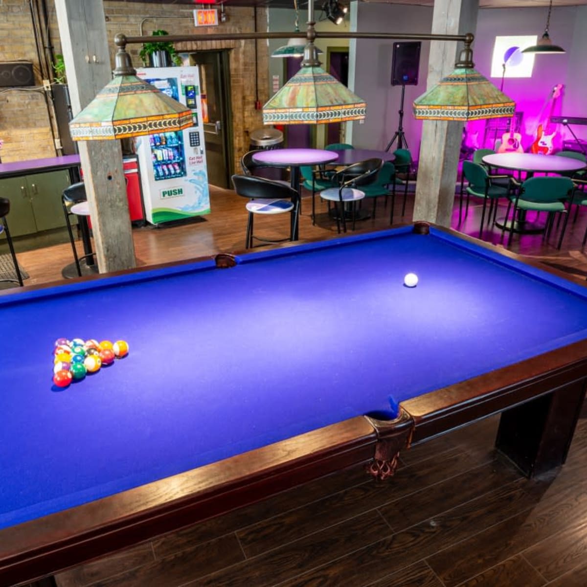 A billiards table with a triangle of colored balls set on its blue felt surface is positioned in a modern recreational room with pendant lighting and lounge seating.