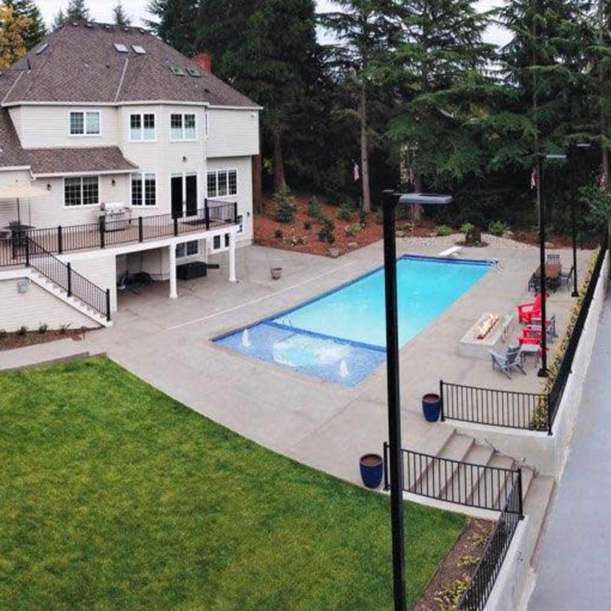 A large two-story house with a swimming pool, a trampoline, and a tennis court in a landscaped yard surrounded by trees.