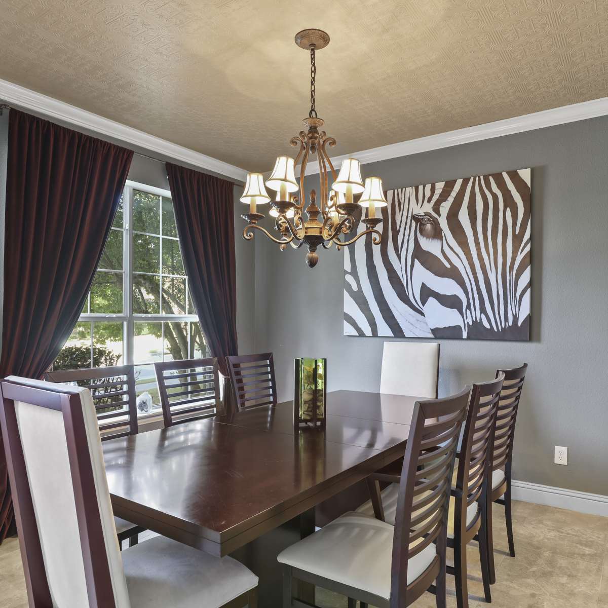 A dining room features a large wooden table surrounded by eight chairs, with a chandelier overhead and a zebra-striped artwork on the wall.