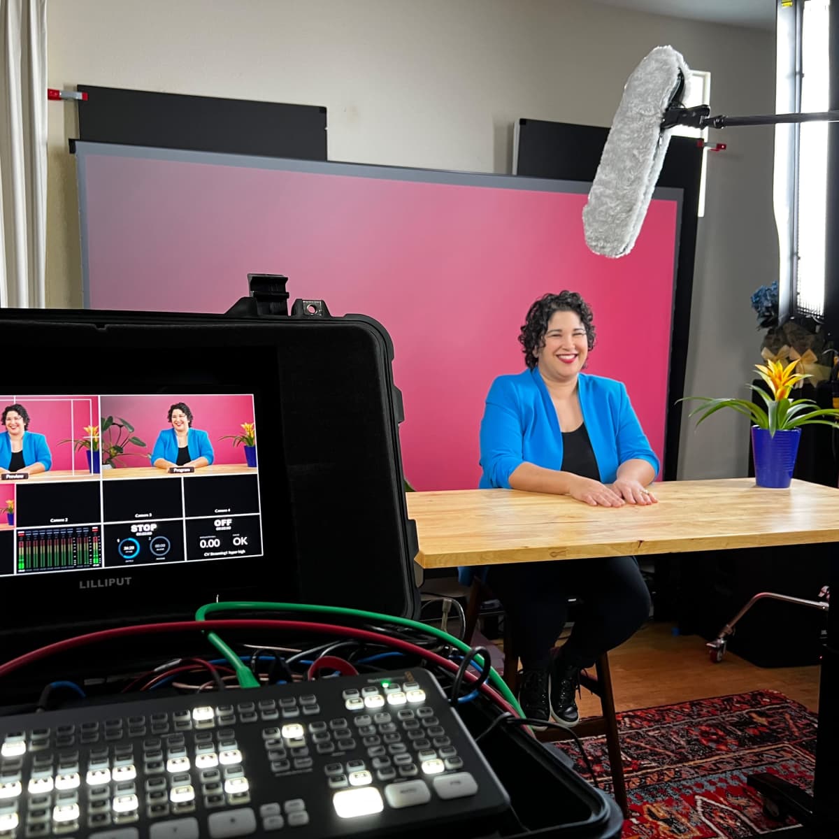 A woman in a blue blazer sits at a wooden table in a studio setup with a pink backdrop and a microphone overhead.
