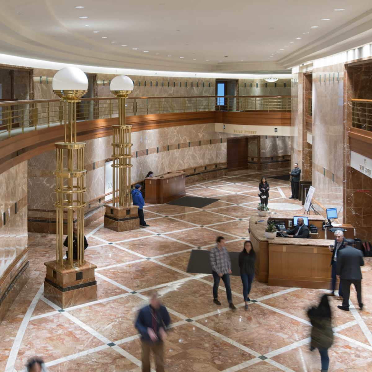 A marble-floored lobby with a curved railing, large light fixtures, and several people walking near the reception desk.